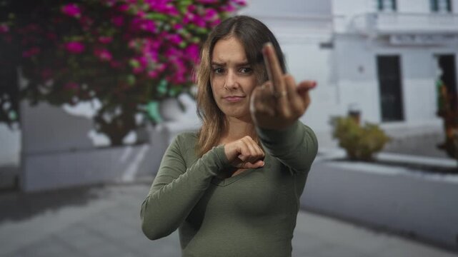 Young hispanic woman shows middle finger to camera in a city street framed by white building and pink bougainvillea; defiance.