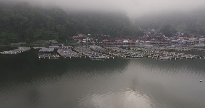 Aerial view of fish farms in a lake near a village shrouded in mist, creating an ethereal scene, Terunyan, Bali, Indonesia.