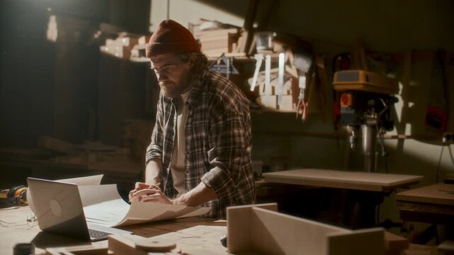Craftsman using laptop and sketching plan on paper at workbench in carpentry workshop, preparing before woodworking project
