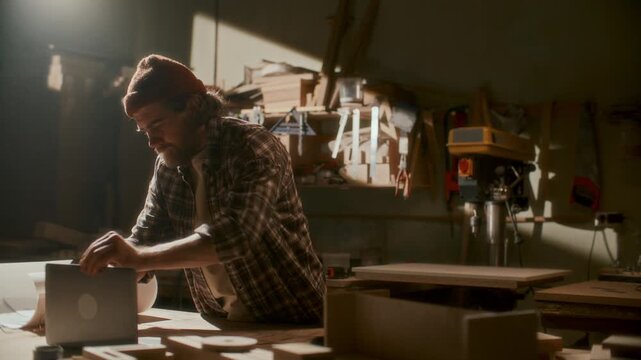 Modern carpenter in plaid shirt and beanie hat opening laptop, reviewing blueprints and surfing the Internet at workbench in warmly lit woodworking workshop
