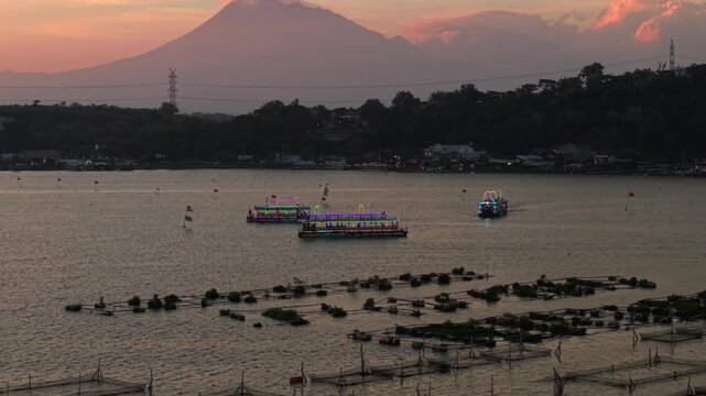 Aerial view of boats with colorful lights sailing on Rowo Jombor Lake against the backdrop of a mountain at sunset, Klaten Regency, Indonesia.