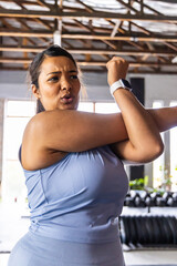 Asian woman stretching upper body inside gym near windows and dumbbells, pulling arm across chest