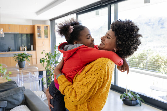Smiling African American mother and daughter embracing near sliding glass doors, with potted plants