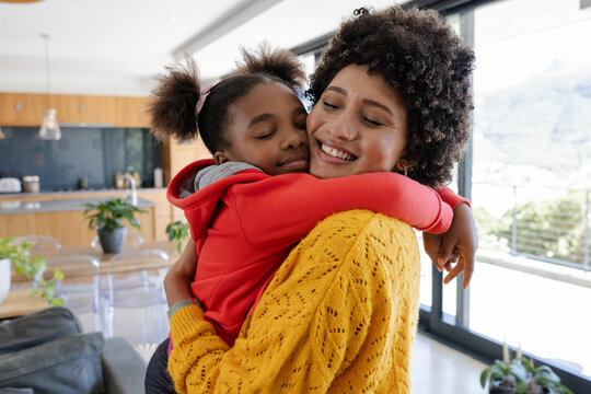 Hugging diverse mother and daughter wearing knit sweater and hoodie in home kitchen, with balcony