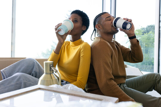 Sitting African American couple drinking from paper cups on couch in living room, with moving boxes