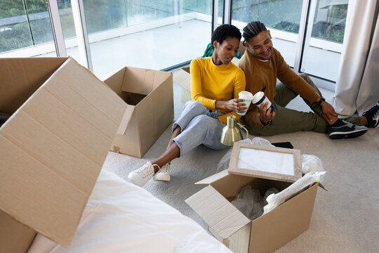 Holding coffee cups, Hispanic and African American couple resting on living room floor, among boxes