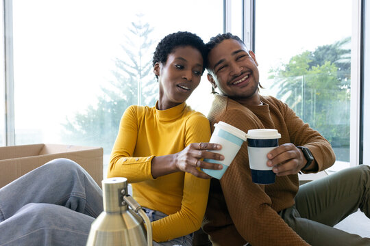 Clinking reusable cups, diverse couple leaning back-to-back in living room, with box and lamp base