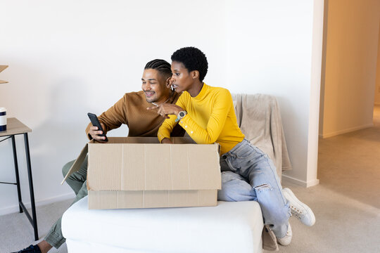 Pointing at smartphone, diverse couple sitting on white ottoman in living area, with cardboard box