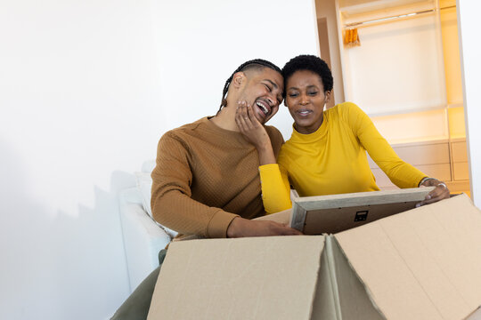 Unpacking framed picture African American woman and man on white sofa at home, with moving box