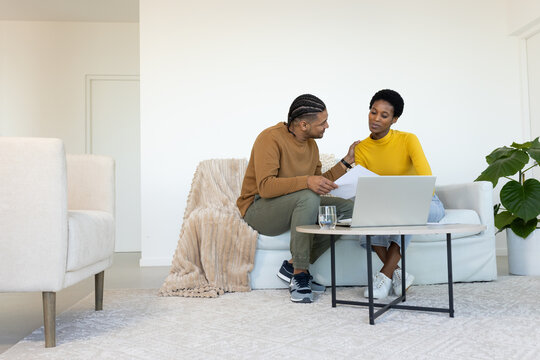 Diverse couple reviewing documents on sofa in living room, with laptop on coffee table