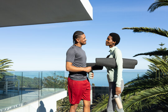 Standing workout partners holding rolled yoga mats and drinks on balcony terrace overlooking ocean