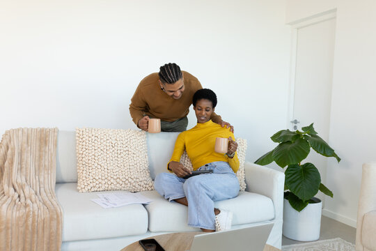 Leaning Diverse couple holding coffee mugs and viewing tablet on sofa at home, with laptop papers