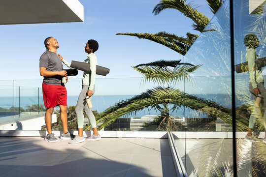 Standing diverse friends holding yoga mats and water bottles on modern terrace, with palm tree view