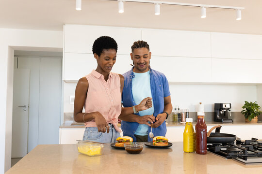 Cooking Diverse couple assembling burgers at quartz kitchen island, with cheese, mustard, ketchup