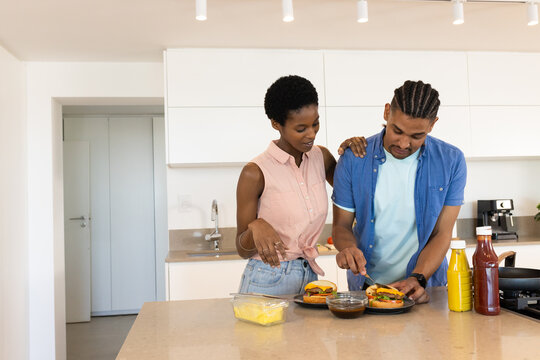 Standing Diverse couple preparing burgers at kitchen island, with ketchup mustard and cheese slices