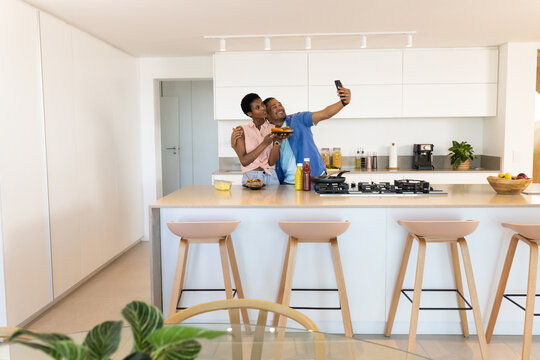Snapping selfie diverse couple standing behind home kitchen island, with smartphone and fruit bowl