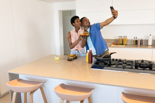 Taking selfie Diverse couple hugging behind kitchen island in modern kitchen, with phone and burger