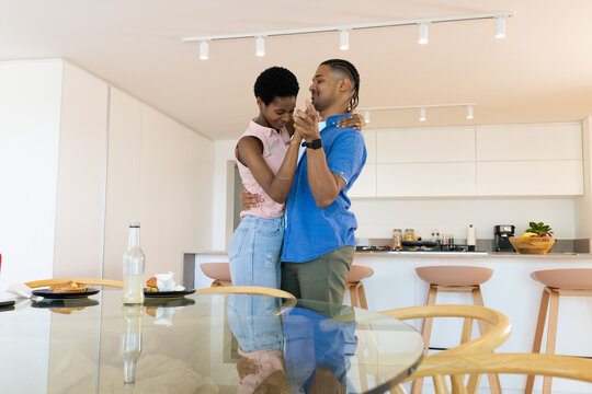 Dancing Diverse couple leaning together in modern kitchen, with glass dining table and fruit bowl