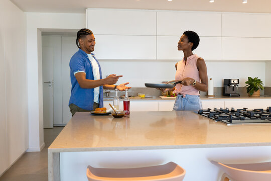 Cooking couple standing at bright kitchen island, with frying pan, hamburger and condiment bottles