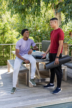 Chatting diverse male friends sitting on bench by table on backyard deck, with rolled yoga mats