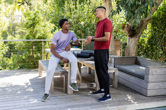 Diverse male friends talking on backyard wooden deck, with rolled-up yoga mats and water bottle