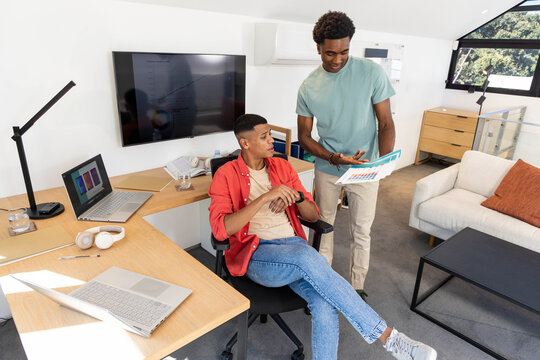 Diverse coworkers reviewing printed chart in office loft, with laptops and monitor