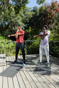 Stretching male friends extending arms across chest on deck, with exercise mats and water bottle