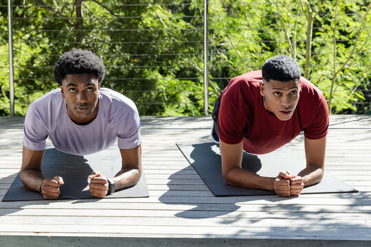 Diverse male friends holding plank position on wooden deck, with exercise mats and smartwatch