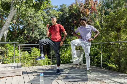 Diverse male friends in sportswear doing knee-raises on backyard deck, with mats and water bottle
