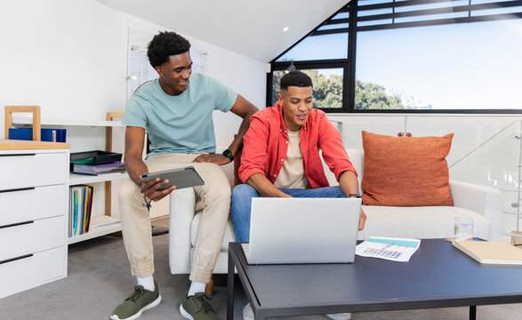 Diverse male friends working on laptop and tablet in loft living room, with papers, coffee table