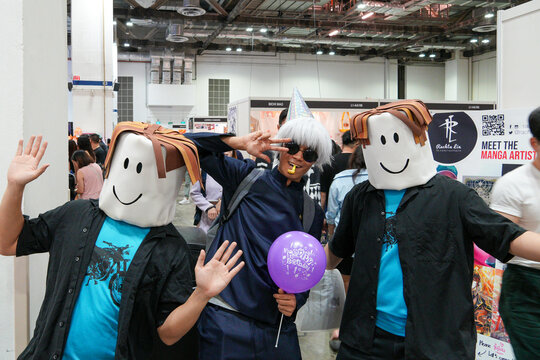 Three people pose with smiling cartoon character masks. One holds a purple balloon. They wave and gesture at a convention event. Bright lights illuminate the indoor space