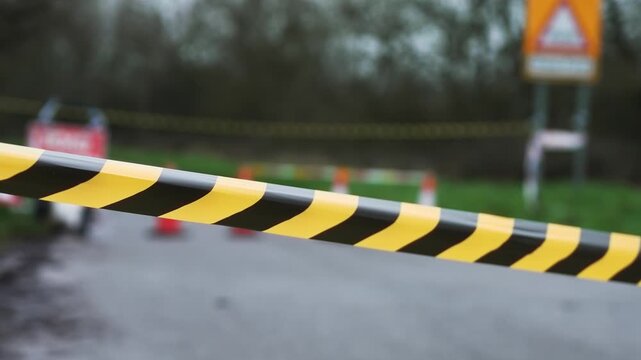 Black and yellow caution tape stretching across restricted roadway. Road Closed sign and traffic cones blocking passage in background. Temporary prohibition restricting vehicle entry and enforcing