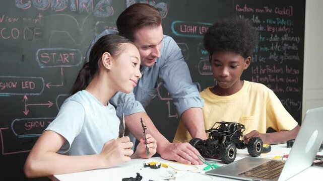 Caucasian teacher pointing at laptop screen while explain car model system at blackboard with coding or programming code written. Happy diverse student learning about robotic construction. Edification