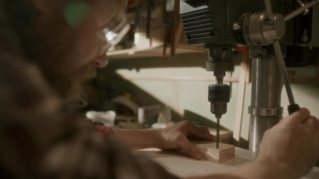 Professional woodworker in protective eyewear boring hole into wooden block with drill press machine, stabilizing material by hand at workbench. Rack focus, close-up shot