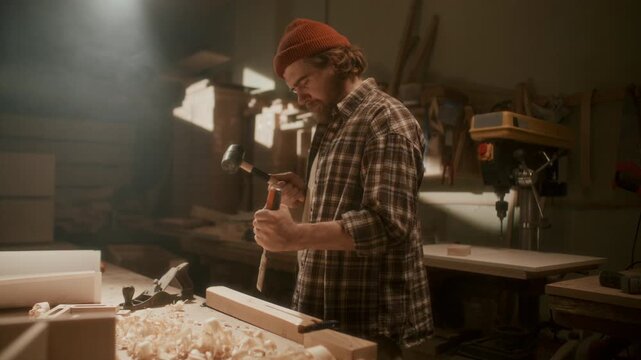 Skilled woodworker carving wooden piece with chisel and mallet in workshop, shaping material with strikes at workbench during carpentry process