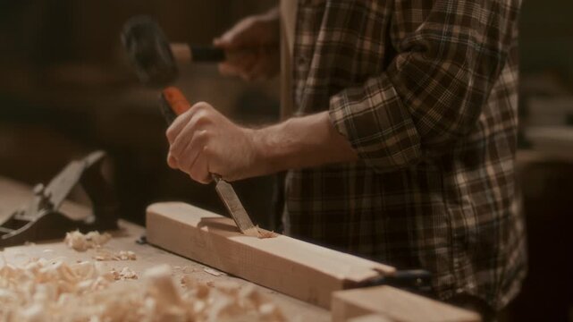 Artisan shaping wooden board with chisel and mallet in workshop, carving material with strikes at workbench in woodworking workshop