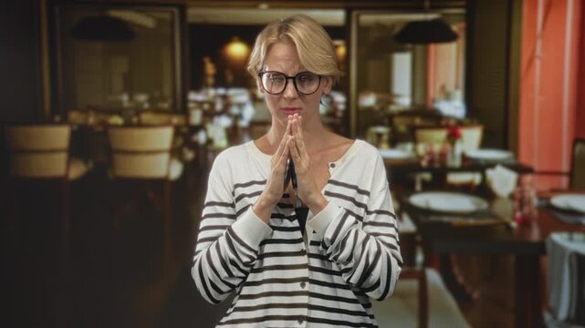 Blond caucasian woman wearing glasses and striped sweater holding car keys with clasped hands in a restaurant dining room; quiet contemplation.