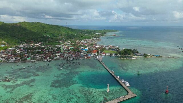 Aerial view of Karimunjawa village, where buildings meet turquoise sea and lush green hills, displaying a contrast of textures and tones, Karimunjawa, Central Java, Indonesia.