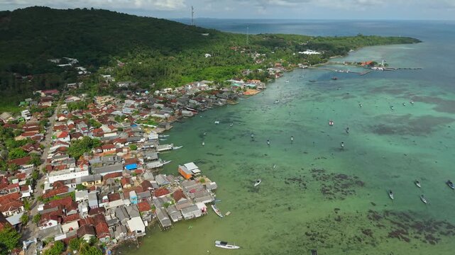 Aerial view of boats dotting the turquoise waters by a densely populated coastal village contrasted against lush green hills, Karimunjawa, Central Java, Indonesia.