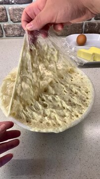 Close-up of a hand peeling off cling film from a bowl of bubbly, risen yeast starter. Showing airy texture and fermentation process of live dough. Professional food photography of home baking steps.