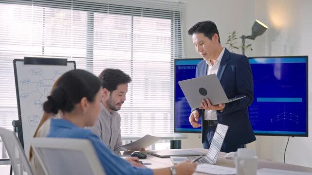 Professional Asian business team meeting discussing project updates and collaborating on laptop during a routine office meeting.