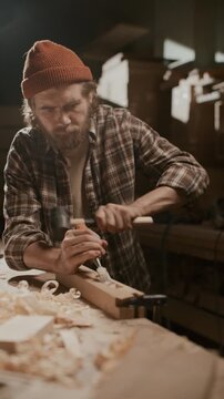 Bearded male carpenter wearing plaid shirt and knit beanie carving mortise into wooden plank using hand chisel and mallet at workbench in joinery workshop. Vertical shot