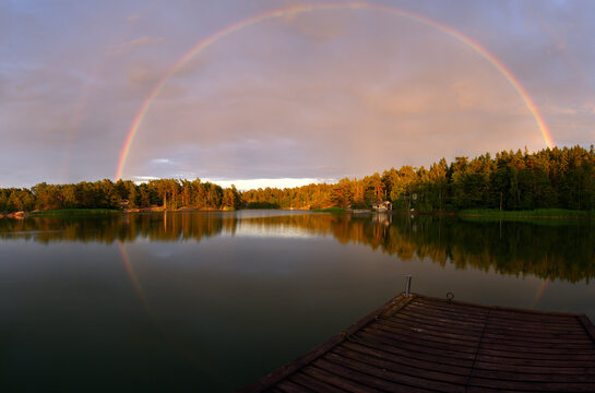 A breathtaking panoramic landscape captures a full rainbow arching across the sky over a calm lake. The rainbow is mirrored in the still water, creating a near-perfect circular reflection.