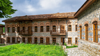 Obraz premium Courtyard of Shaki Khans Mosque Complex in Shaki, Azerbaijan. Historic stone and brick building with wooden balconies, a tiled roof, and green grass under a cloudy sky