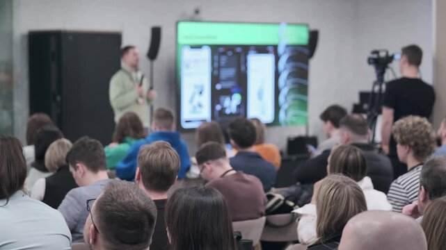 Business tech conference presentation, speaker addresses engaged audience in modern conference room, attendees listen and take notes, camera records session.