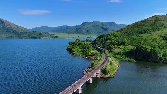 A captivating high-angle shot of a freight train crossing a bridge over the Bukhtarma Reservoir. The train travels along the scenic shoreline surrounded by lush green trees and bushes. A bright, sunny