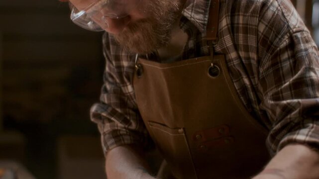 Woodworker in safety goggles and earmuffs finishing wooden plank with electric belt sander, smoothing surface as sawdust and shavings dispersing across workshop bench. Tilt down shot