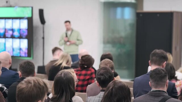 Attendees listen to presenter at tech conference, seated audience facing stage, networking atmosphere, modern meeting room.