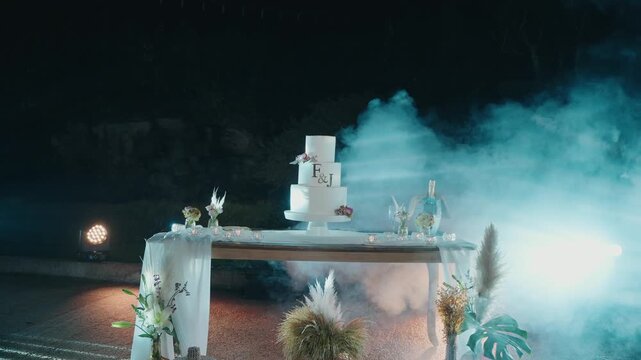 White tiered wedding cake with monogram on draped table surrounded by smoke and blue light