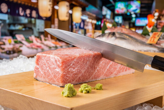 Sharp Japanese knife resting on a premium block of fresh raw fatty tuna with wasabi on a wooden board at a bustling authentic Asian seafood market.